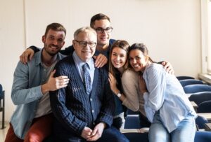 Una figura de experiencia y mentoría, un pilar relevante en la consultoría empresarial, posa con un grupo de jóvenes profesionales. La imagen irradia un ambiente de confianza y colaboración entre las generaciones, demostrando la importancia de la guía y el conocimiento compartido para el futuro de cualquier negocio.