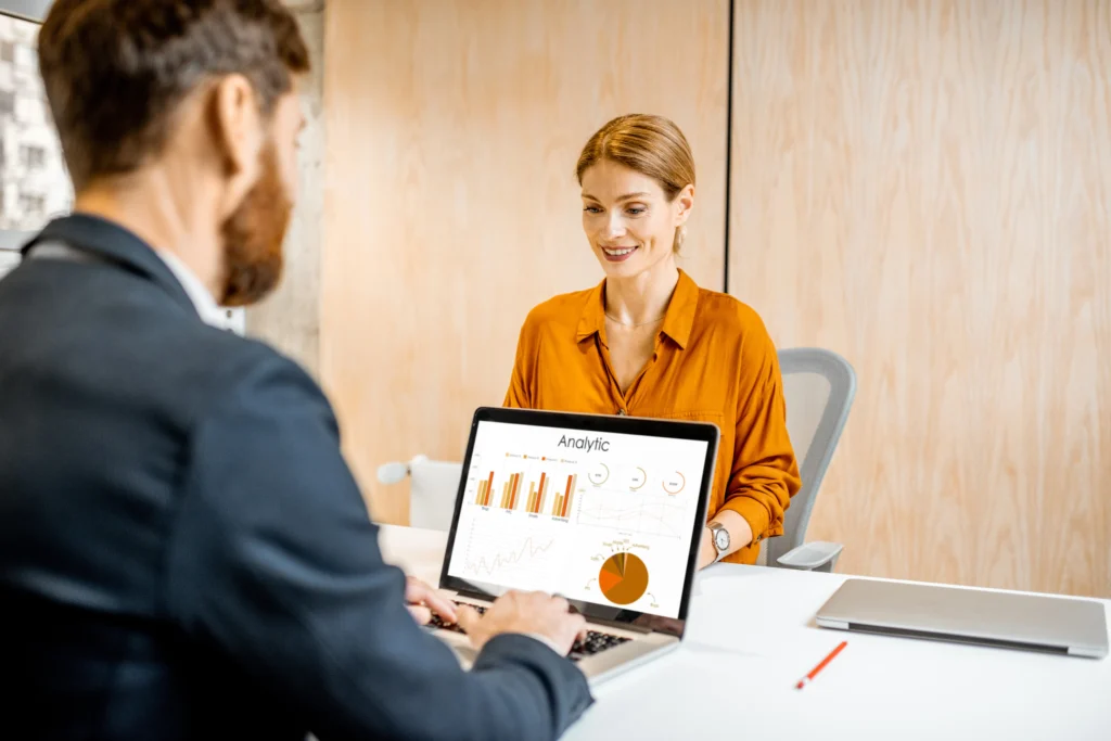 Una profesional observa mientras un colega muestra análisis de datos en un laptop, relacionando el rendimiento con el perfil de puestos.